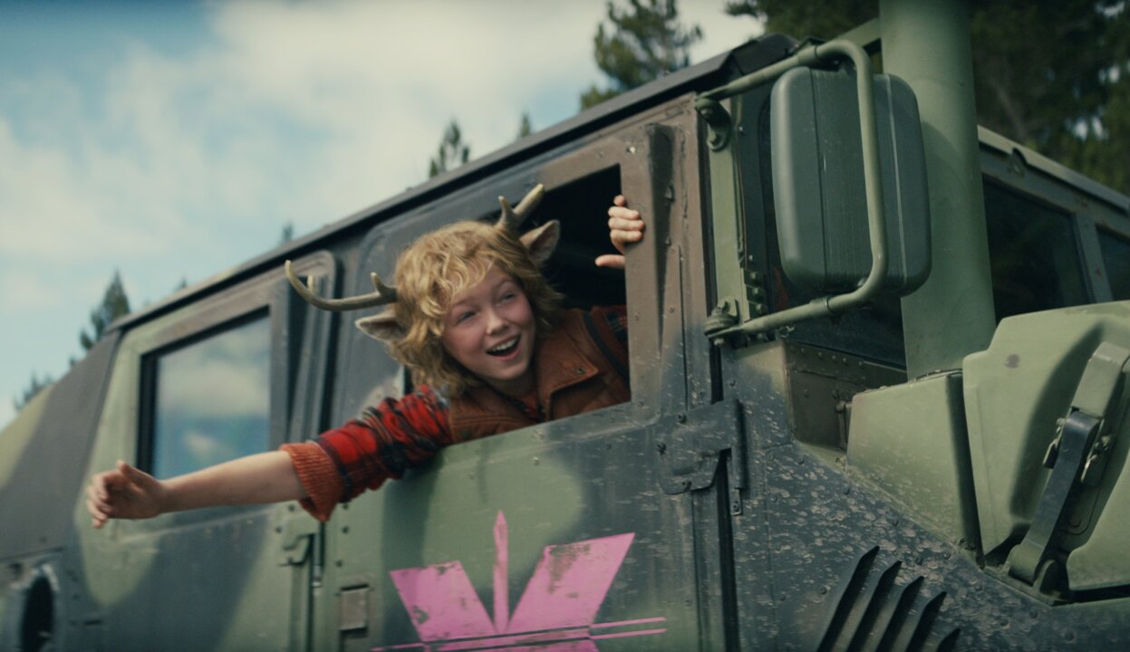 Gus, de Sweet Tooth, um garoto com chifres de cervo, sorridente dentro de um veículo acenando na janela.